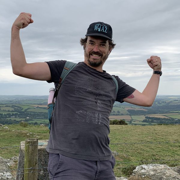 Gabriel Stebbing on a hilltop in a cap and glasses, grinning and flexing both arms